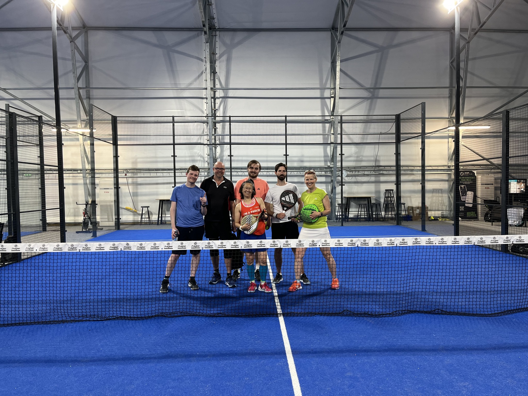 Gruppering av fyra killar och två tjejer på en padelbana bakom nät i en padellokal.
