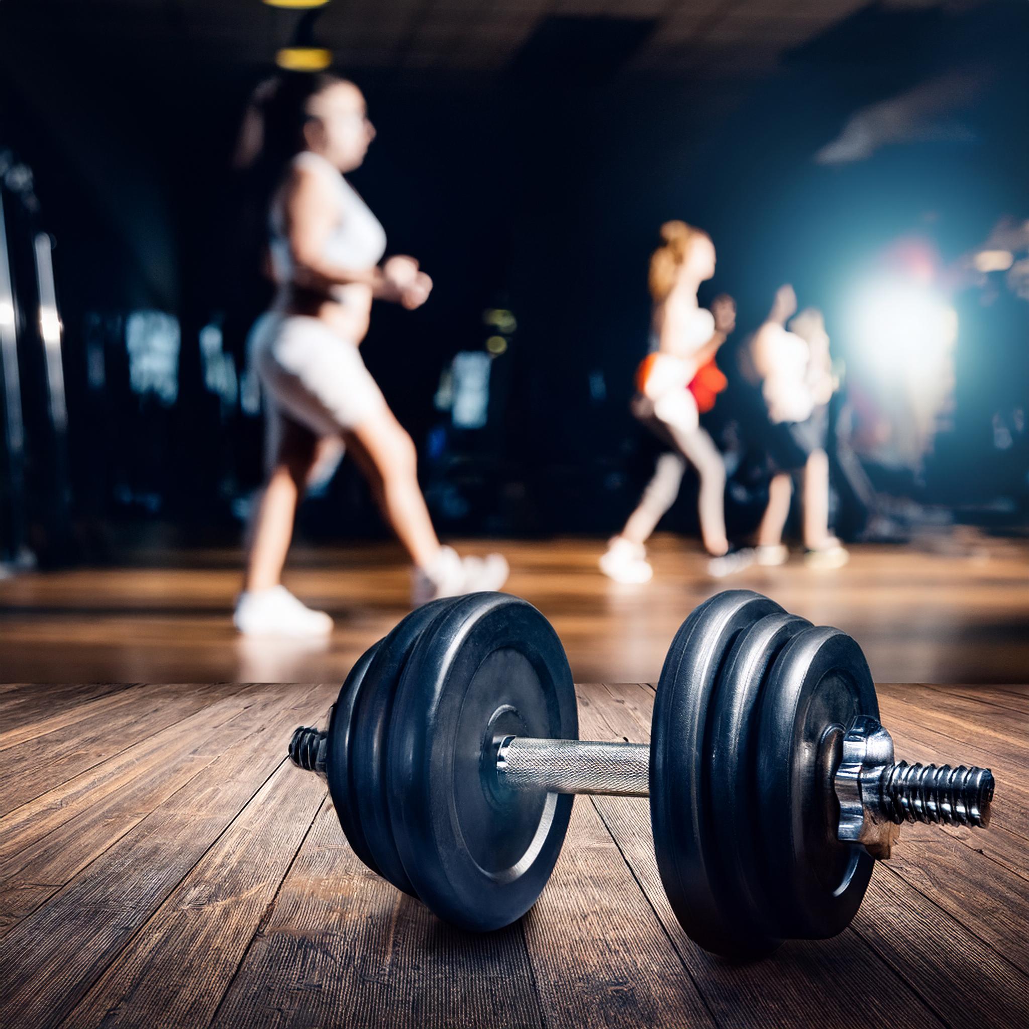 en gymlokal med en hantel i förgrunden på att trägolv. gymdeltagare i bakgrunden som tränar genom att hålla i vikter. Lokalen är lite mörk och stämningsfull.
