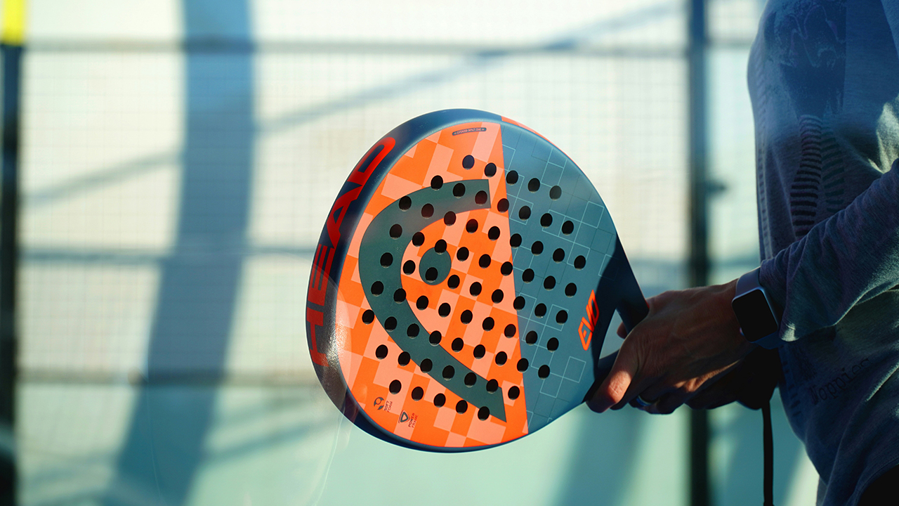 En person håller i ett padelrack som är orange/grått på en padelbana i kvällsljus.